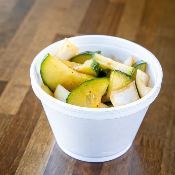 A white styrofoam cup filled with Hawaiian pickled mango and cucumber on a wooden table.